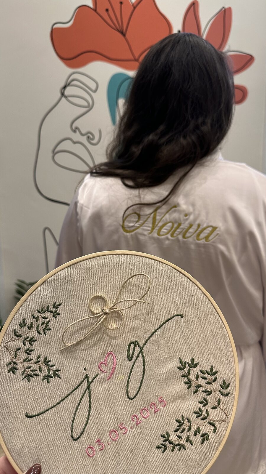 Noiva de costas com robe bordado 'Noiva' segurando bastidor com iniciais do casal e data do casamento em ponto-cruz — Salão do Bosque