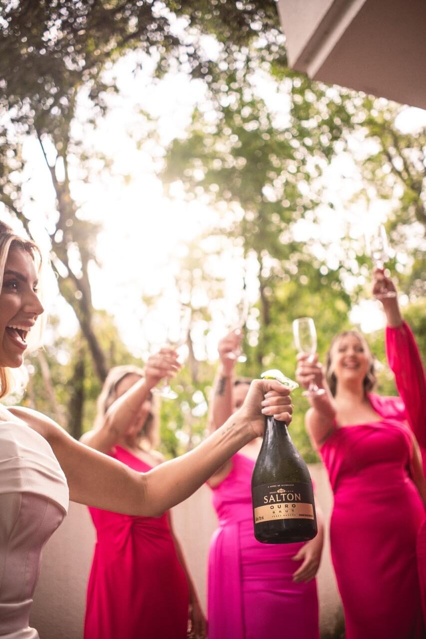 Noiva abrindo champanhe com madrinhas em vestidos rosé brindando no jardim